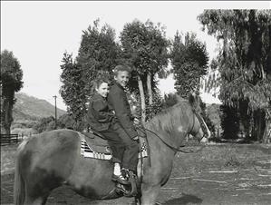 Two children seated together on the back of a brown horse with palm trees in the background