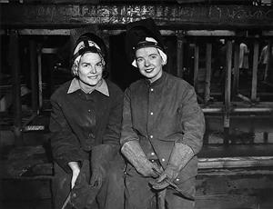 Two white women in work suits, gloves, and helmets, are sitting and smiling at the camera with dark steel structures behind them