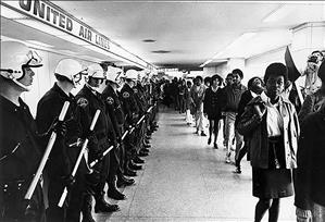 A long row of white police with batons and helmets face a long row of Black men and women inside an airport
