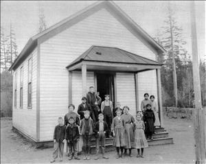 A group of White children and teacher beside three Native Americans in front of a one room schoolhouse