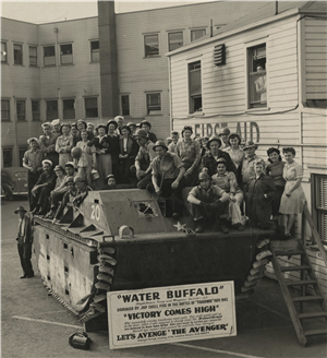 A large group of young men and women sit on top of a military tank in front of a three story building
