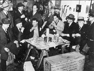 Thirteen men in overcoats, suits and ties sitting on crates, gathered around a small work table smiling and eating