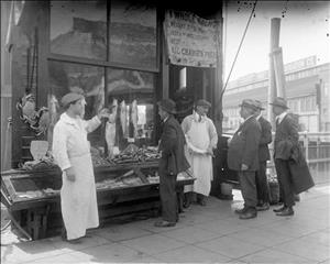 Two fishmongers outside of a storefront holding salmon for men in suits and hats with a cloth sign hanging above the doorway with illegible prices for seafood