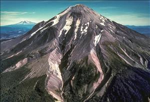 An aerial view of a volcano with very little snow on top. In the background are snowy peaks and a blue sky