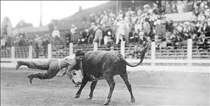 Rider with both feet in the air being flung by a bull as a crowd watches in bleachers in the background