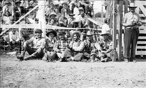 Six men sit against a wire fence smiling in hats and flannel shirts in front of the seated rodeo audience