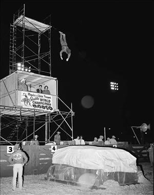 Man jumping from scaffolding raised above an announcer's booth onto an object covered in tarp as men in cowboy hats look on