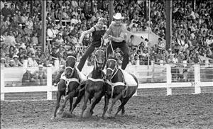 Two riders standing on and holding the reigns of three racing horses as a crowd looks on in the bleachers