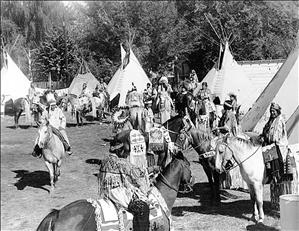 A large group of Indigenous people in ceremonial dress beside five conical tent structures