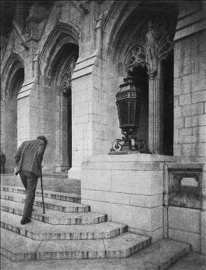 Exterior shot of a man climbing a set of stone stairs with a cane towards a gothic building with a large, ornate lantern at the entrance