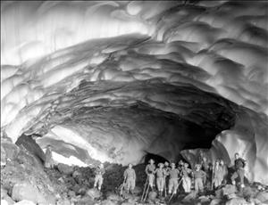 Fourteen people in outdoor wear with hats and walking canes pose inside of am arena sized ice cave covered in smooth, contoured divots