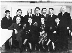 A group of eleven people pose next to a piano on a hardwood floor in formal clothing