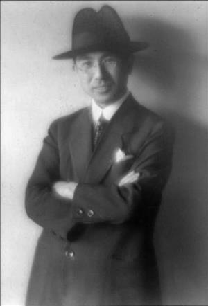 Person in a brimmed hat, round glasses and suit and tie smiling against a wall