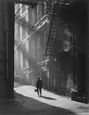 Man walking into an alleyway lined with dramatically lit fire escapes and electrical lines