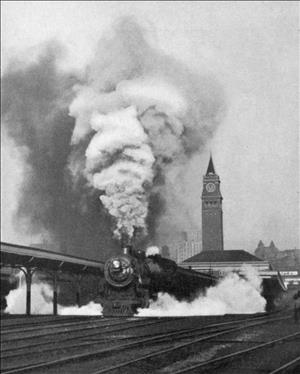 A train pouring out steam leaving the station with a brick clock tower in the distance