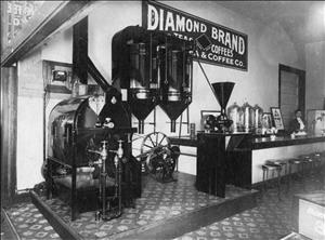 Interior of coffee shop with roasting equipment in front of a bar with an attendant behind the counter, where there is a sign reading Diamond Brand Teas and Coffees"