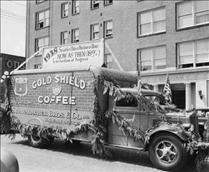 A delivery truck decorated with conifer tree limbs and American flags, with signage reading 1938, Seattle's Oldest Business House, Now as then (1897) In the Forefront of Progress, Gold Shield Coffee"