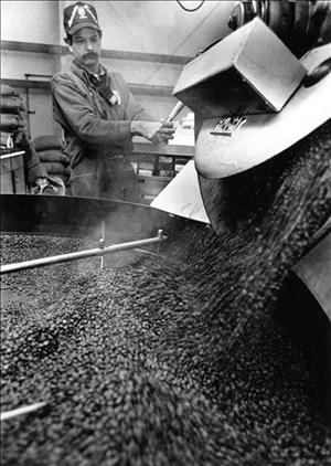 A worker in a baseball hat and jumpsuit pours steaming coffee beans into a mixer