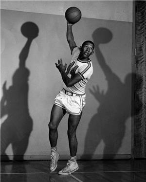 A basketball player in uniform in a hook shot pose casting two distinct shadows behind him on the gymnasium wall