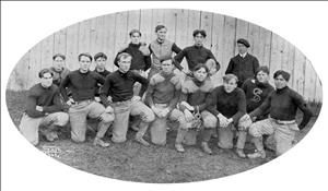 Thirteen football players kneeling and standing in long sleeve black wool shirts, striped pants lined with cushioned knee and shoulder pads beside the coach, in front of wood panels